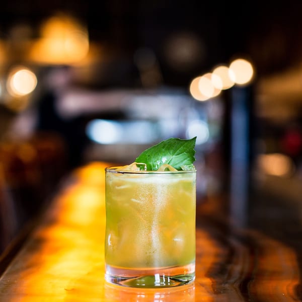 A vibrant green cocktail with ice and a basil leaf garnish on a glowing bar top.