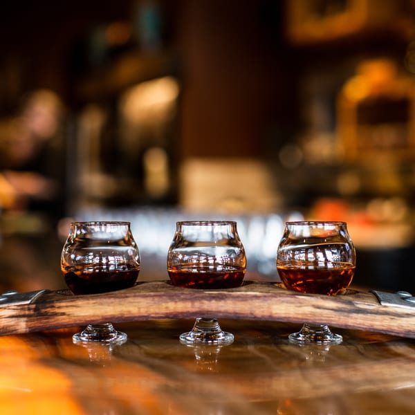 Three glasses of amber liquid, possibly whiskey, on a wooden barrel at Unwined Kitchen & Bar.