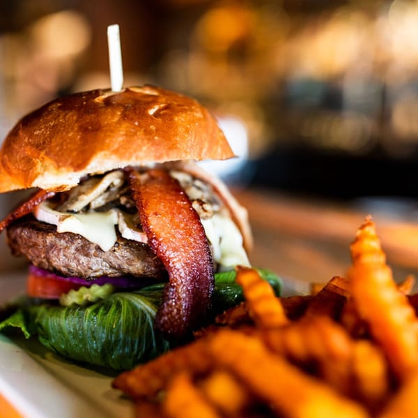 Gourmet burger with bacon, mushrooms, cheese, and vegetables, served with sweet potato fries.