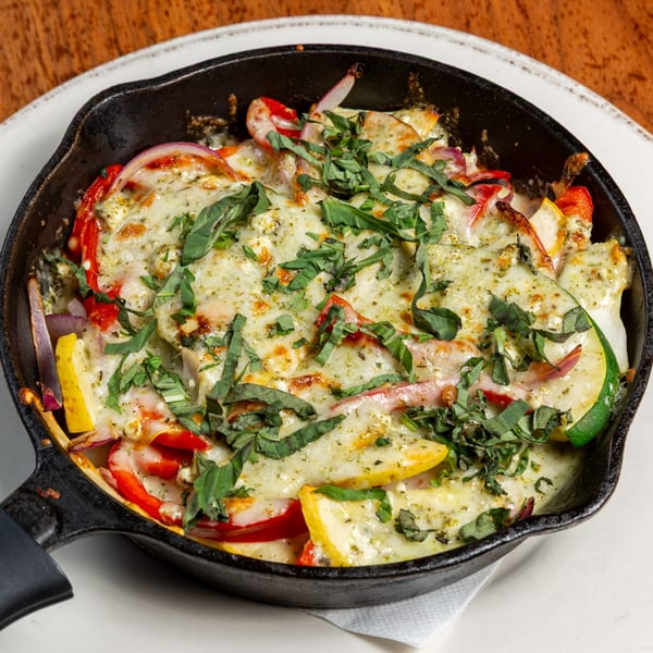 Skillet of melted cheese over sliced zucchini, red peppers, and onions, garnished with fresh basil.
