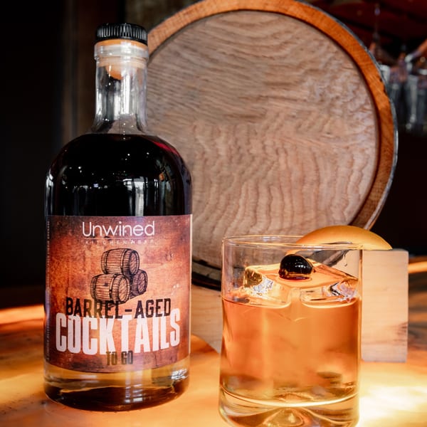 Unwined Kitchen & Bar barrel-aged cocktail bottle and a glass with ice, cherry, and orange peel.