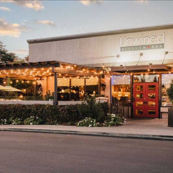 Exterior of Unwined Kitchen & Bar with a lit patio, red doors, and lush landscaping.