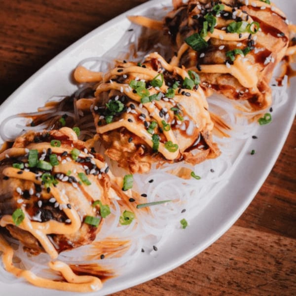 Three fried dumplings drizzled with orange sauce, sesame seeds, and green onions on a bed of clear noodles.